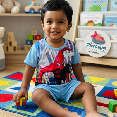 Criança sorridente vestindo camisola azul claro com Homem-Aranha, sentada num tapete colorido com blocos de madeira e brinquedos ao fundo.