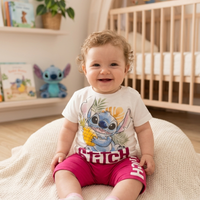 Bebé sorridente com roupa estampada de Stitch sentado em manta bege dentro de quarto com berço de madeira