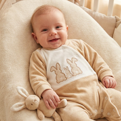 Bebé sorridente de macacão bege e branco com aplicação de coelhos, deitado numa almofada bege tricotada e segurando peluche coelho.