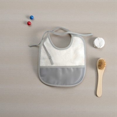 Babete branco com bolso cinzento e fita para atar rodeado por escova de madeira, disco de algodão e bolas azul e vermelha numa superfície bege.