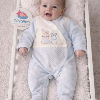 Bebé em macacão azul claro deitado em cama branca com etiqueta Pirralhos