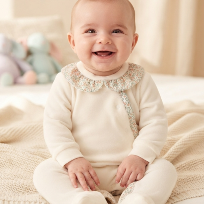 Bebé sorridente com macacão creme e gola floral sentado numa cama clara