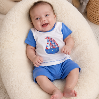 Bebé sorridente com conjunto branco e azul com barco, sentado em almofadão creme felpudo.