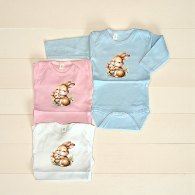 Três bodies de bebé com desenho de lebres abraçadas em cores azul, rosa e branco sobre fundo branco.