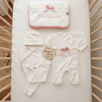 Conjunto de roupa de bebé branco com detalhes rosa num berço de madeira clara