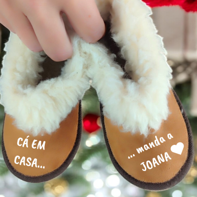 Pantufas castanhas forradas com pele sintética branca e texto nas solas, seguradas por mão, fundo desfocado com luzes.