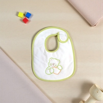 Babete branco com borda verde claro e bordado de ursinho colorido sobre superfície clara ao lado de cubos coloridos