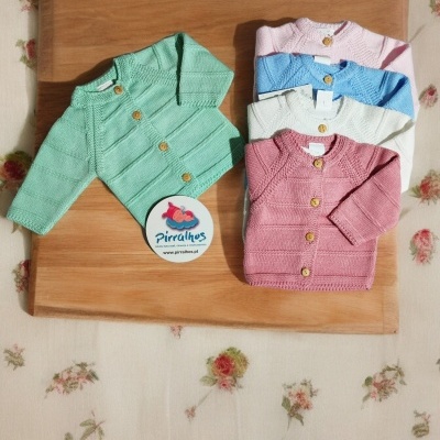 Casacos de bebé coloridos em malha com botões de madeira sobre tábua de madeira e fundo floral.
