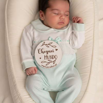 Bebé a dormir em almofada branca com macacão verde claro às riscas e branco com colarinho e laço, segurando placa de madeira com texto