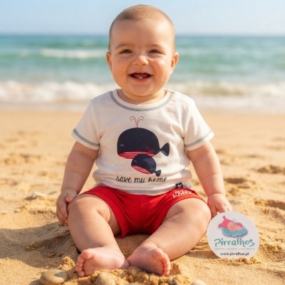 Bebé na praia com roupa branca e vermelha, segurando cartão redondo Pirralhos