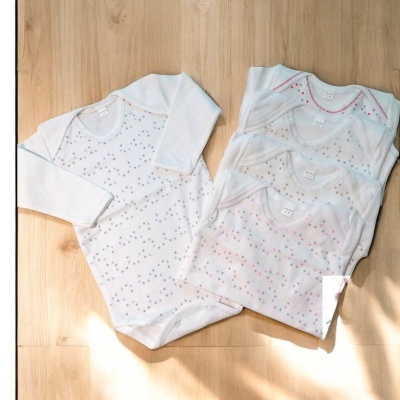 Bodies de bebé brancos com padrão de pontos coloridos em cruz sobre chão de madeira clara