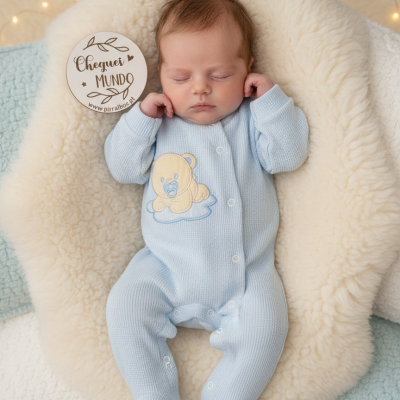 Bebé adormecido em fato de pijama azul claro com ursinho bordado numa almofada felpuda creme