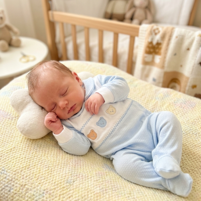 Bebé dormindo em pijama azul com ursinhos, deitado em almofada pastel, com berço e peluches ao fundo.