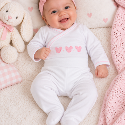Bebé com pijama branco e faixa rosa, deitado com peluche e mantas rosas e creme.