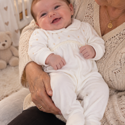 Bebé sorridente com babygrow branco com estrelas douradas nas mãos de mulher com camisola bege de malha.