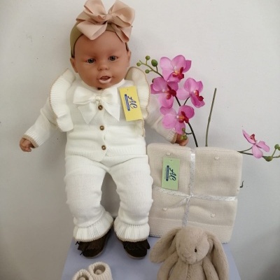 Boneca bebé de malha branca com laço rosa, manta, botinhas e coelho de peluche junto a uma orquídea rosa.