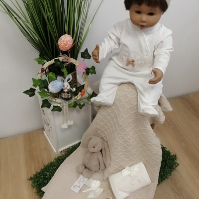 Boneco bebé vestido de branco com laço bege, manta bege texturada, peluche coelho bege, sapatinhos, oferta embrulhada, cesta com decoração e plantas artificiais num fundo branco.