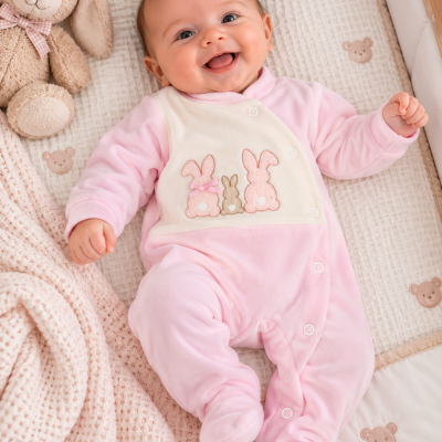Bebé com pijama rosa com coelhinhos bordados deitado em berço acolchoado com manta bege e peluche de coelho