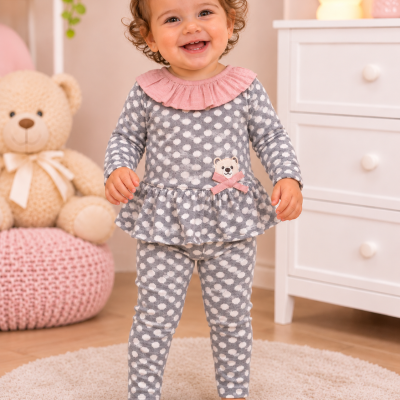 Bebé com roupa cinzenta de bolinhas brancas e gola rosa, de pé em tapete com móveis e brinquedos ao fundo.