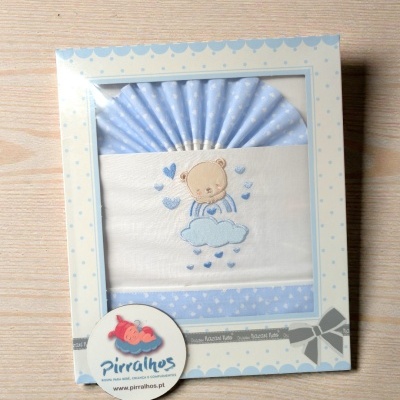 Conjunto para bebé azul com ursinho e nuvem em embalagem branca com pontos azuis sobre superfície de madeira.