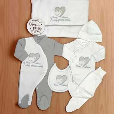 Conjunto de roupa branca e cinzenta para bebé com mensagens bordadas, com vários acessórios e placa de madeira decorativa.