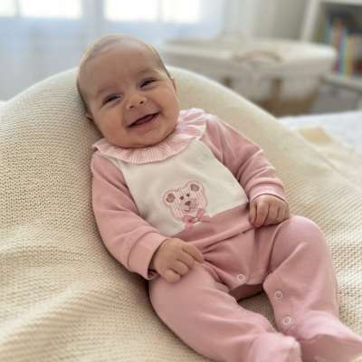 Bebé sorridente de macacão rosa e branco com ursinho bordado na parte frontal, deitado sobre manta bege tricotada.