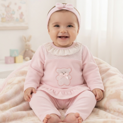 Bebé sorridente com roupa rosa com ursinho bordado sentado em manta creme e rosa.