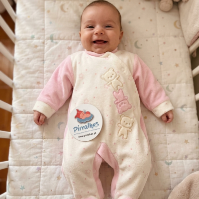 Bebé sorridente de pijama rosa e branco com ursos, deitado em berço acolchoado com padrão de estrelas e luas.