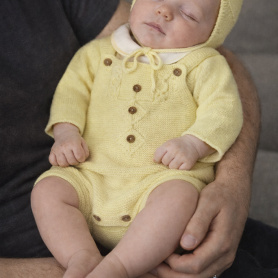 Bebé com macacão e capuz amarelos dormindo no colo de adulto com camiseta cinza escuro