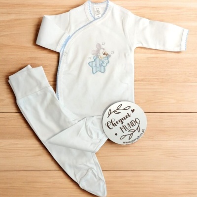 Pijama de bebé branco com ursinho numa estrela azul, fecho lateral e detalhes azuis, sobre madeira clara, com plaqueta redonda branca.