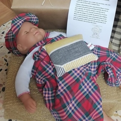 Boneco com roupa xadrez vermelha deitado sobre almofada dourada com papel em português ao lado