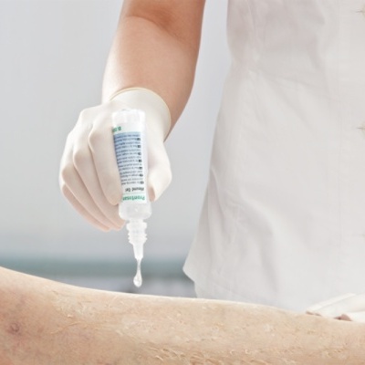 Pessoa com bata branca a aplicar líquido com conta-gotas na perna de um paciente