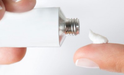 Mão segurando tubo de creme prateado e creme branco no dedo indicador