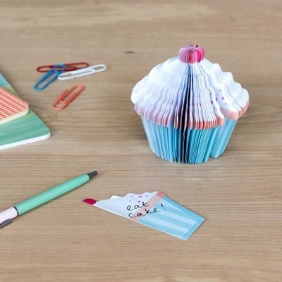 Bloco de papel em forma de cupcake azul, branco e rosa numa mesa de madeira com clipes, caneta e nota escrita 'eat cake!'