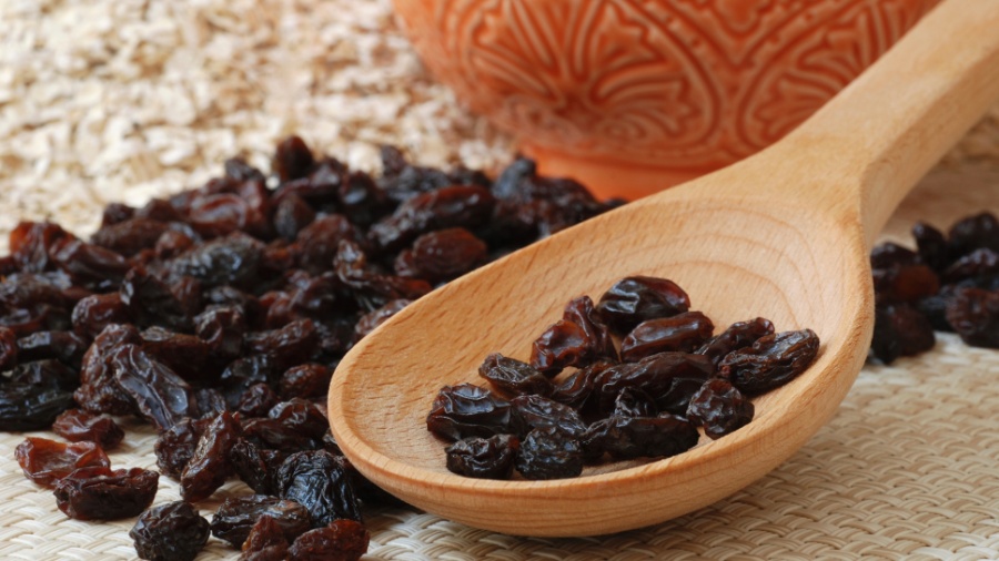 Passas escuras na colher de madeira sobre superfície têxtil bege e vaso laranja ao fundo