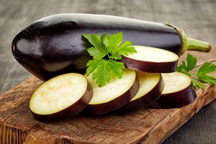 Beringela inteira e fatias sobre tábua de madeira com folhas verdes