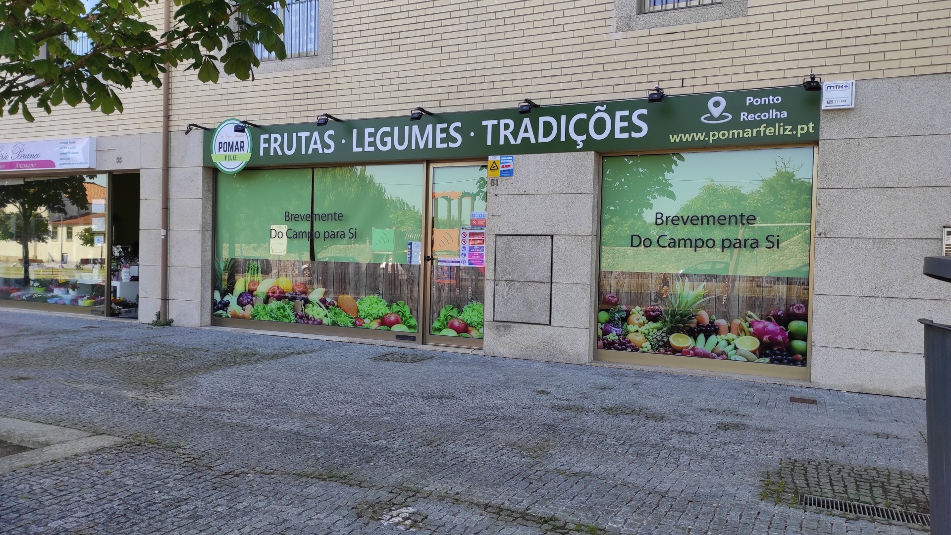 Fachada de loja de frutas e legumes com letreiro azul e janelas decoradas com imagens e texto promocional