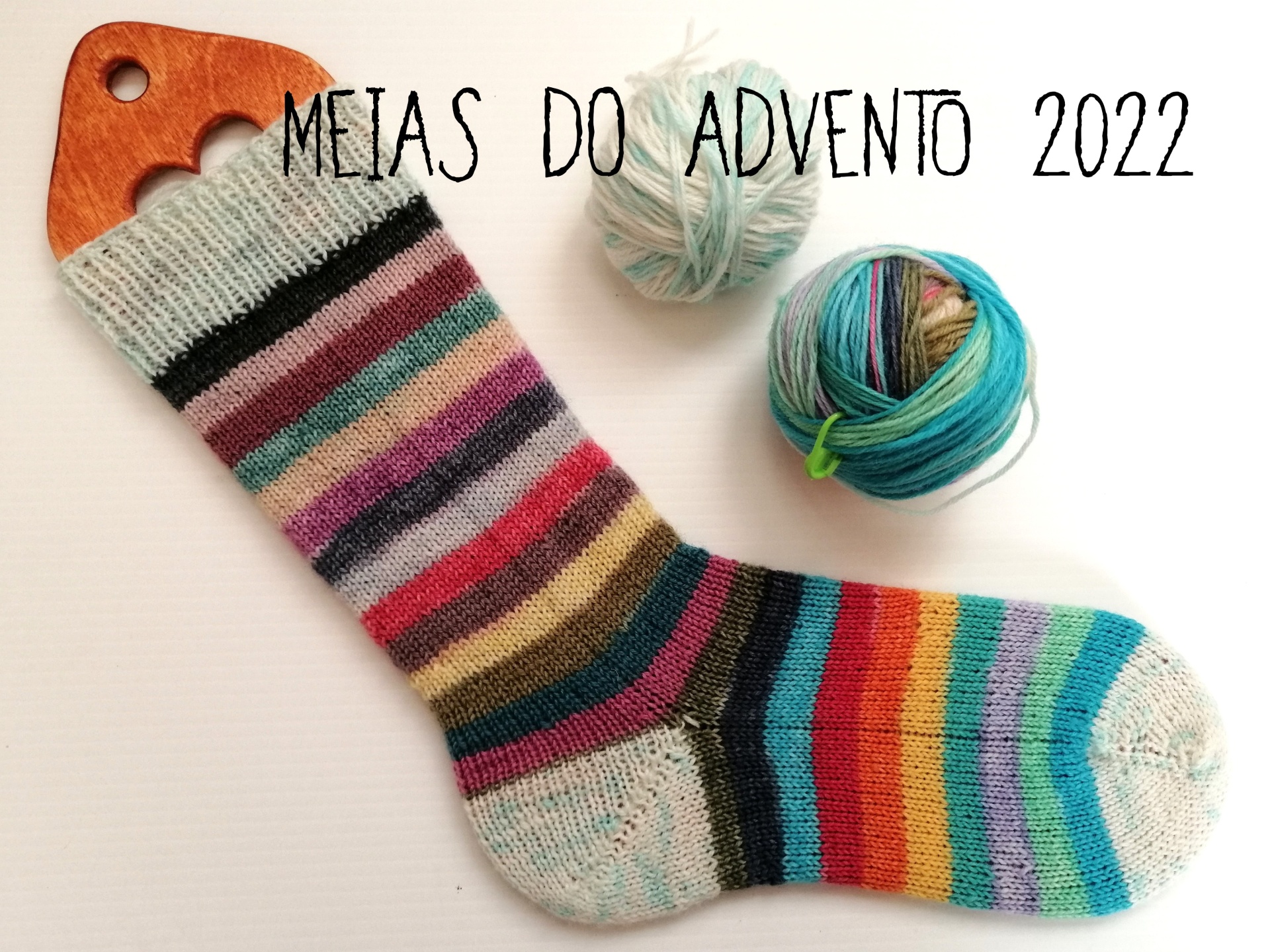 MEIAS DO ADVENTO Meia riscada colorida pendurada num cabide de madeira com duas bolas de lã ao lado