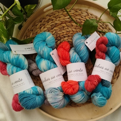 Novelos de lã azul, branco, cinzento e vermelho com etiquetas 'linhos ao vento' numa bandeja de madeira