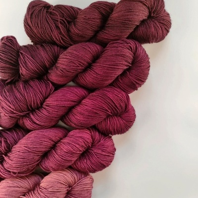 Oito novelos de fio de lã em tons de vinho e rosa dispostos em superfície branca