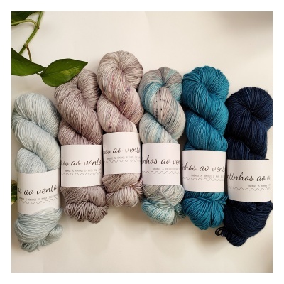 Seis novelos de lã em tons de azul e neutros com etiquetas brancas etiquetadas 'linhos ao vento' sobre mesa clara com planta verde.