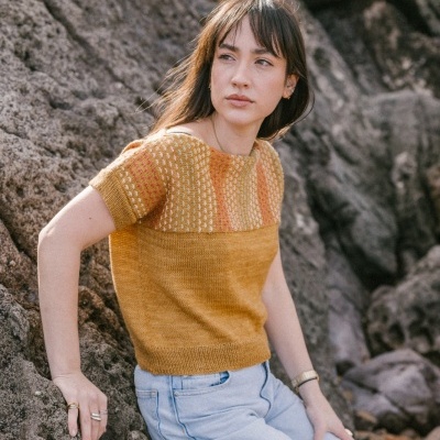 Camisola curta de malha amarelo e laranja com padrão texturizado, mulher posa sentada em rochas, calças jeans claras