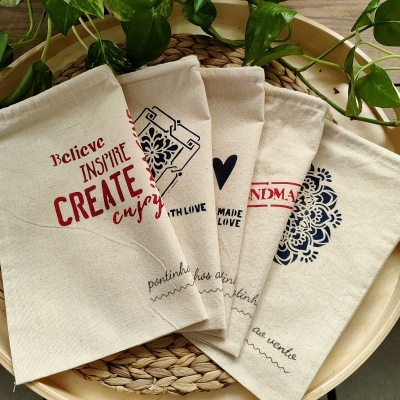 Sacolas de tecido bege com frases motivacionais e padrões florais em vermelho e azul sobre prato de madeira com plantas ao redor