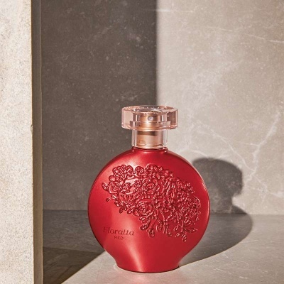 Frasco de perfume vermelho com decoração floral em relevo e tampa transparente sobre superfície de pedra cinza
