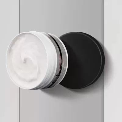 Creme em pote plástico branco com tampa preta e etiqueta cinza sobre superfície cinza clara