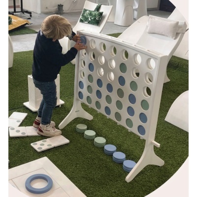 Jogo gigante de ligar quatro em plástico branco com peças azuis e verdes num ambiente interior com relva artificial e cadeiras.