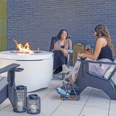 Mesa com lareira integrada a gás e duas mulheres sentadas em cadeiras exteriores, parede de tijolos cinzentos atrás.