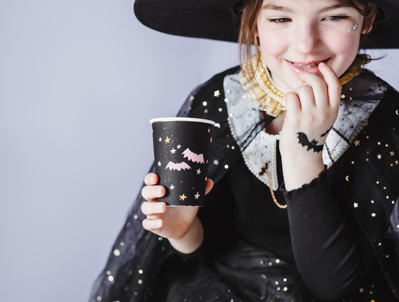 Menina vestida de bruxa segurando copo preto decorado com morcegos e estrelas