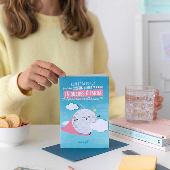 Cartão azul com bola espelhada e capa de super-herói sobre mesa com bolachas e copo de água