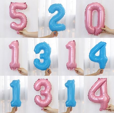 Balões metálicos rosa e azul em formato de números, segurados por mãos contra cortinas brancas.